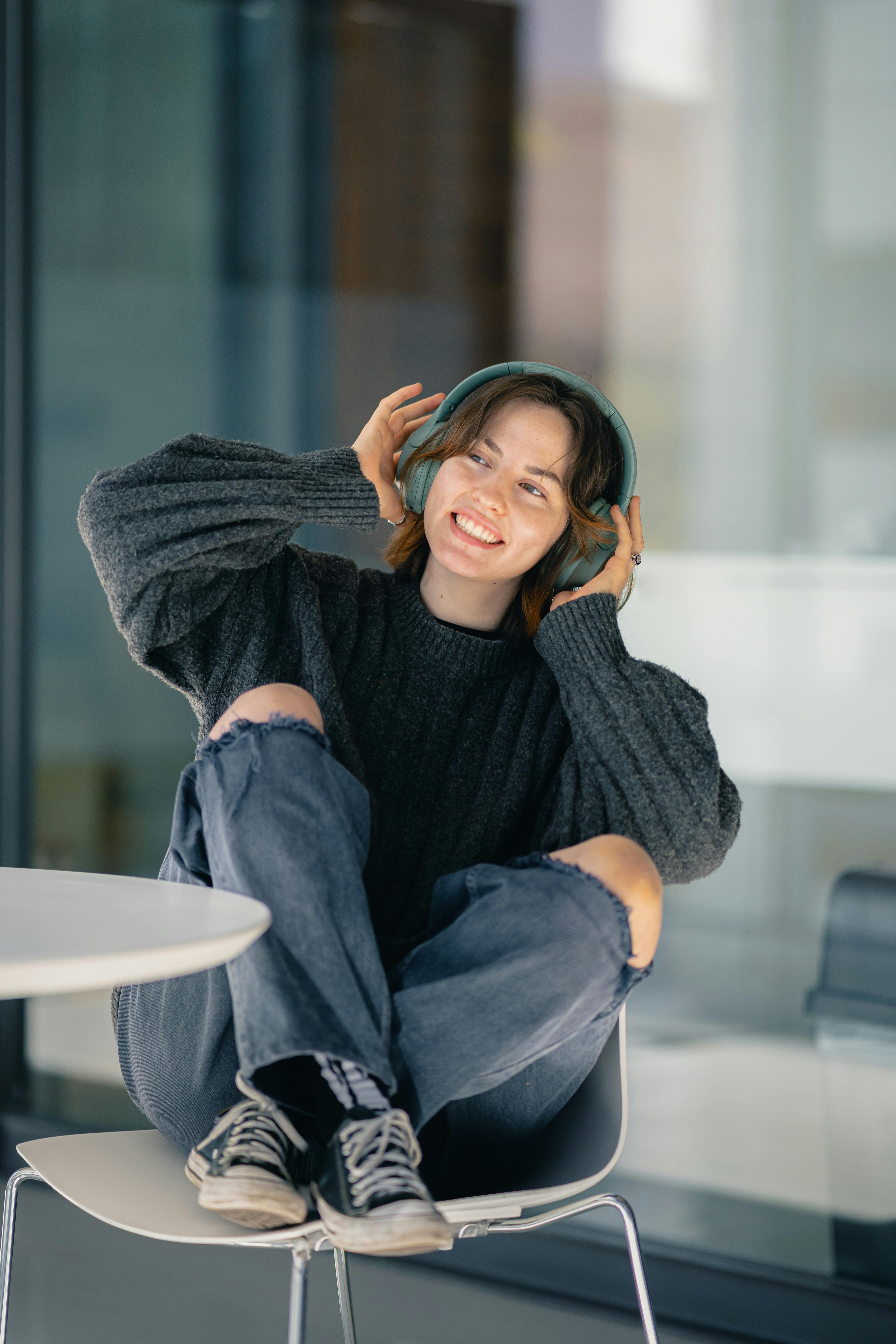 Smiling young woman in casual attire listening to music with headphones indoors.