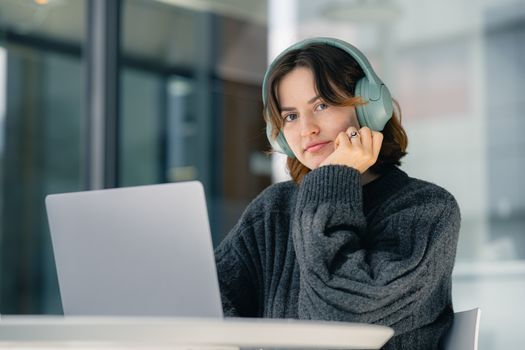 A young woman wearing headphones works on a laptop in a modern indoor setting.