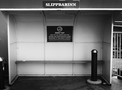 Black and white photo of a smoking shelter in Reykjavik with a humorous sign. Suitable for urban lifestyle themes.