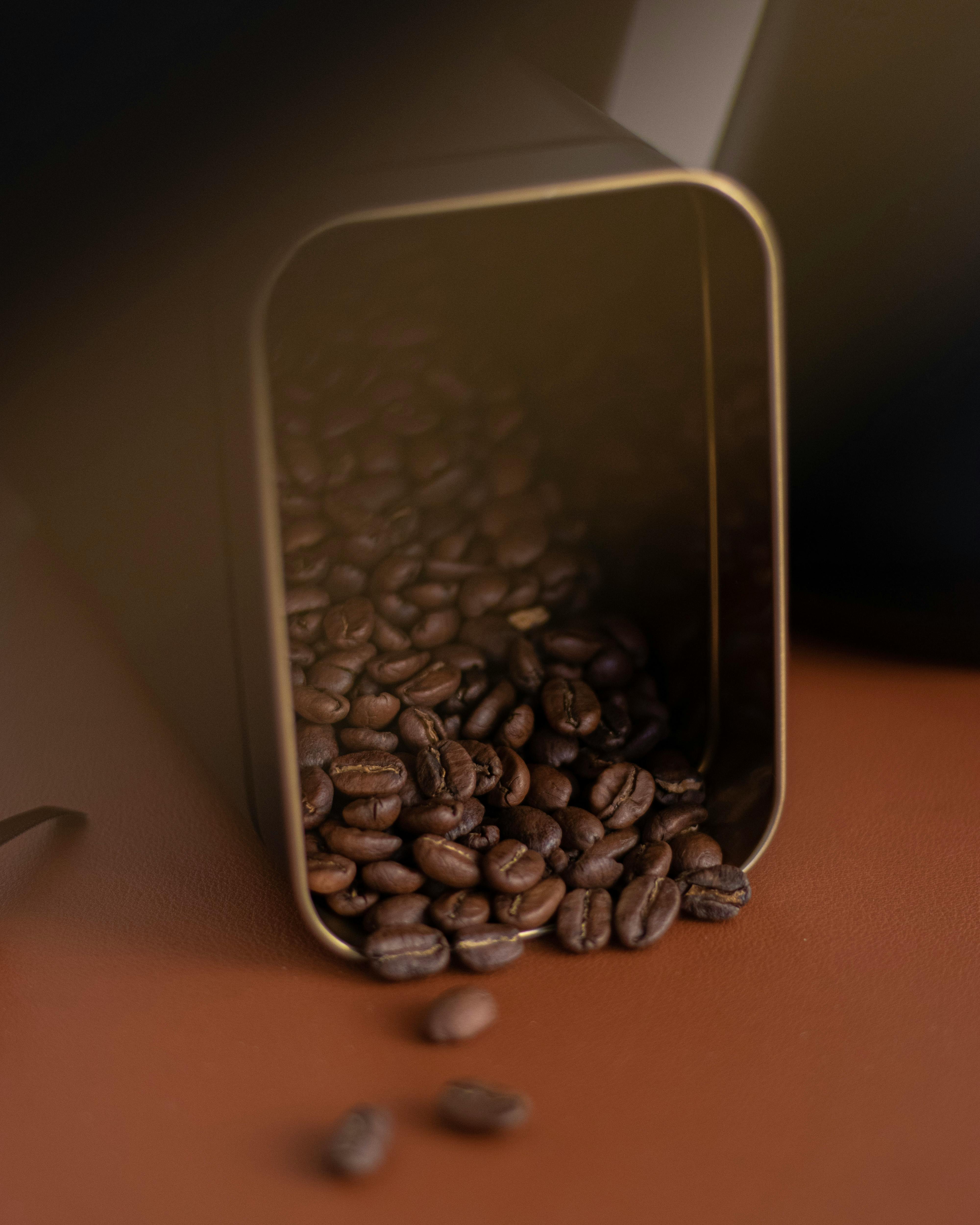 A pile of roasted coffee beans spilling from a square container onto a brown surface.