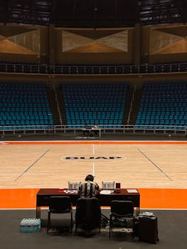 A lone official at a table on an empty basketball court with empty blue seats.