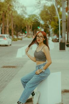Trendy young woman sits on a block in a city street, wearing casual fashion and glasses.
