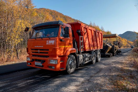 Orange truck and paver working on road construction in an autumn forest with vibrant fall foliage.