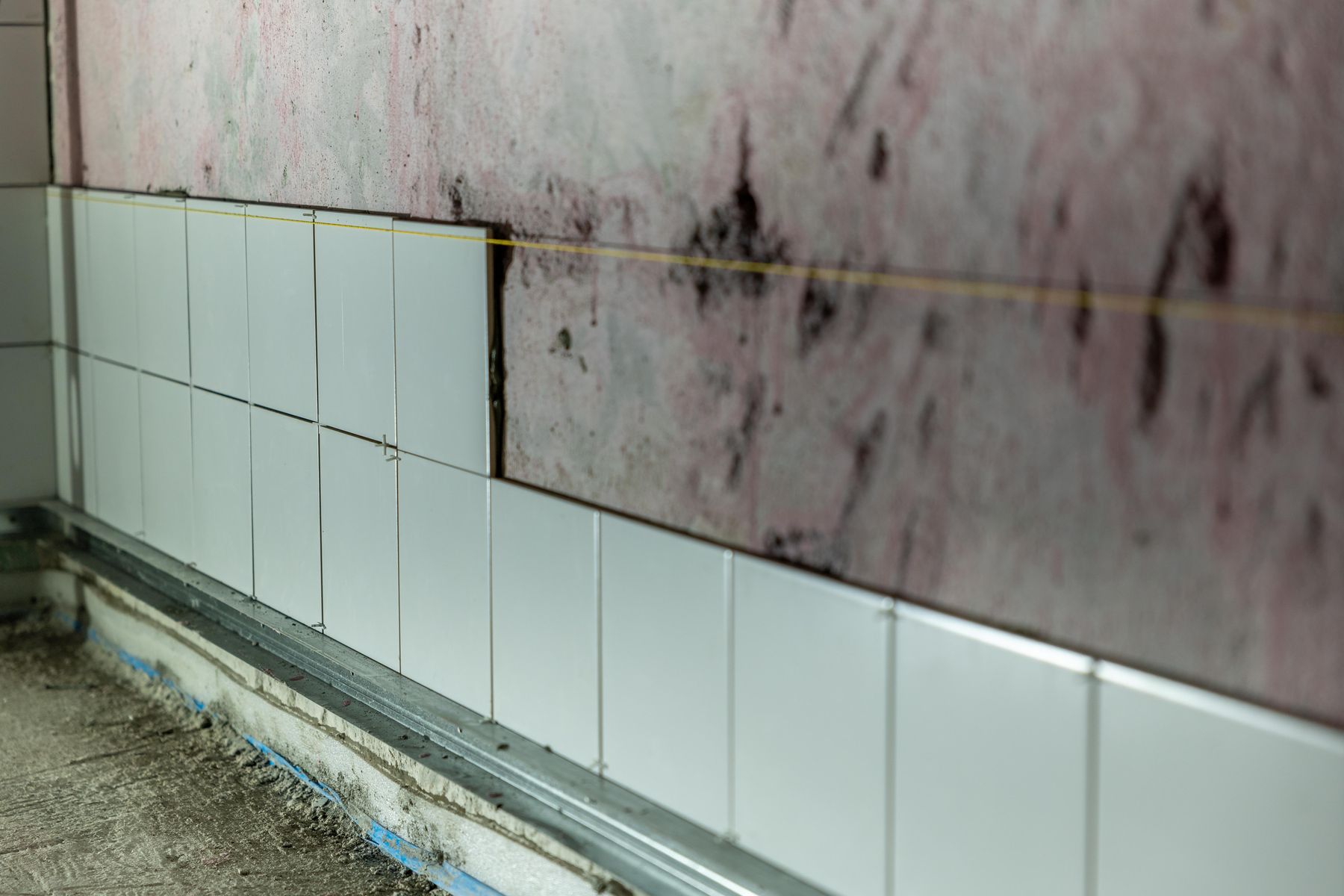 Close-up of a partially tiled wall, showing white wall tiles, exposed substrate, uneven edges and a damp-stained unfinished surface above.