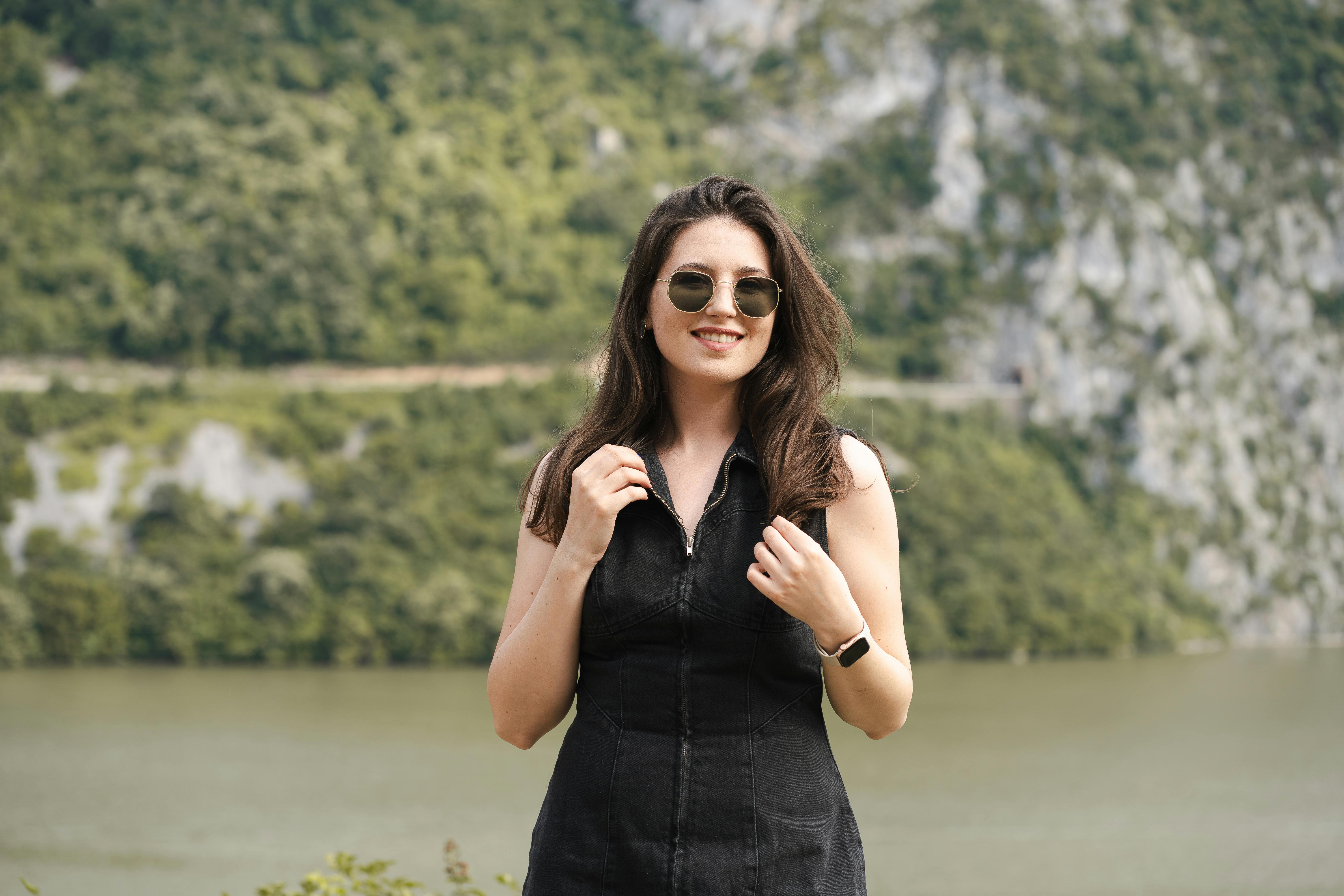 bezplatná Módní žena ve slunečních brýlích s úsměvem u malebného jezera s horami v pozadí. Základová fotografie