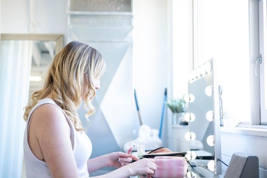 A woman applying makeup at a vanity table with natural lighting in a studio.