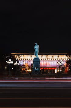 Statue illuminated at night with blurred traffic lights in a historic city setting.