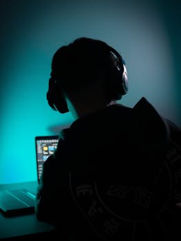 Silhouette of individual wearing headphones, illuminated by screen light, working on a laptop.
