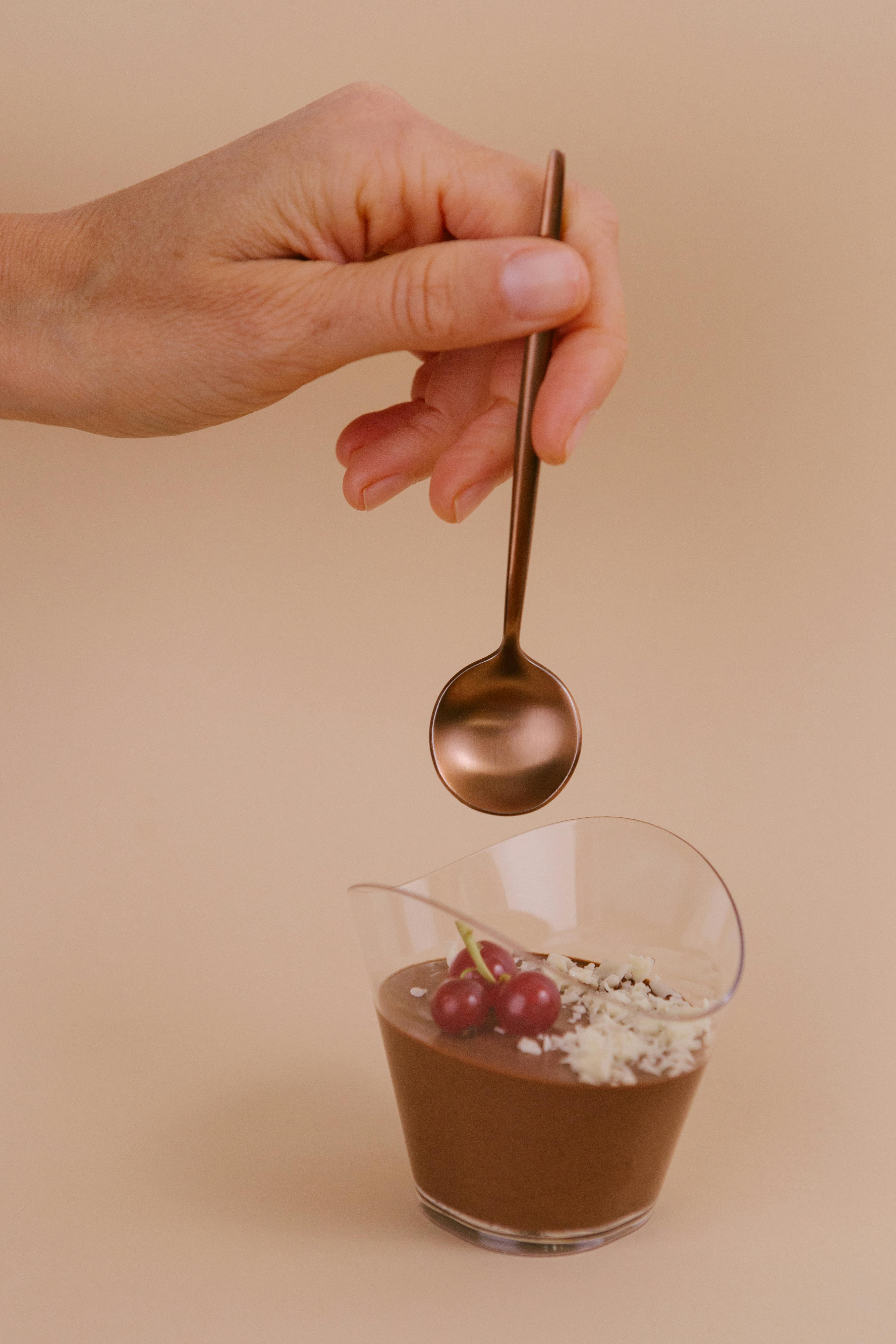 Hand placing spoon over chocolate dessert cup with cherries on a neutral background.