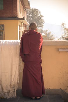 一位身着红色长袍的和尚背对镜头，站在阳光明媚的户外阳台上