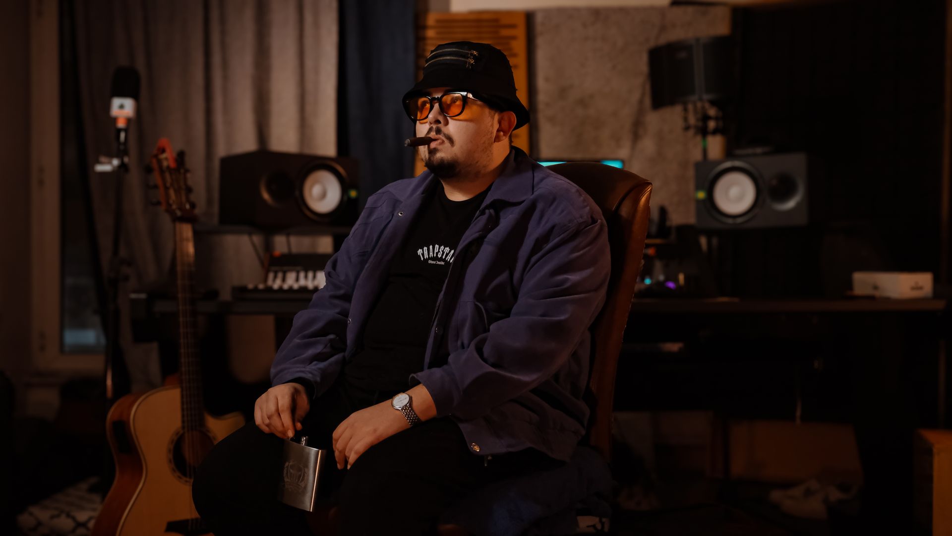 A man sits in a music studio holding a flask, surrounded by guitars and recording equipment.