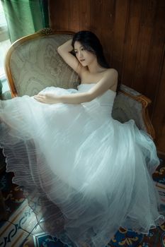 A woman in a white dress resting on a vintage chair, bathed in soft indoor light.