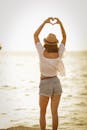 日落时分，一位穿着短裤、戴着帽子的女人在海滩上用双手比出心形
