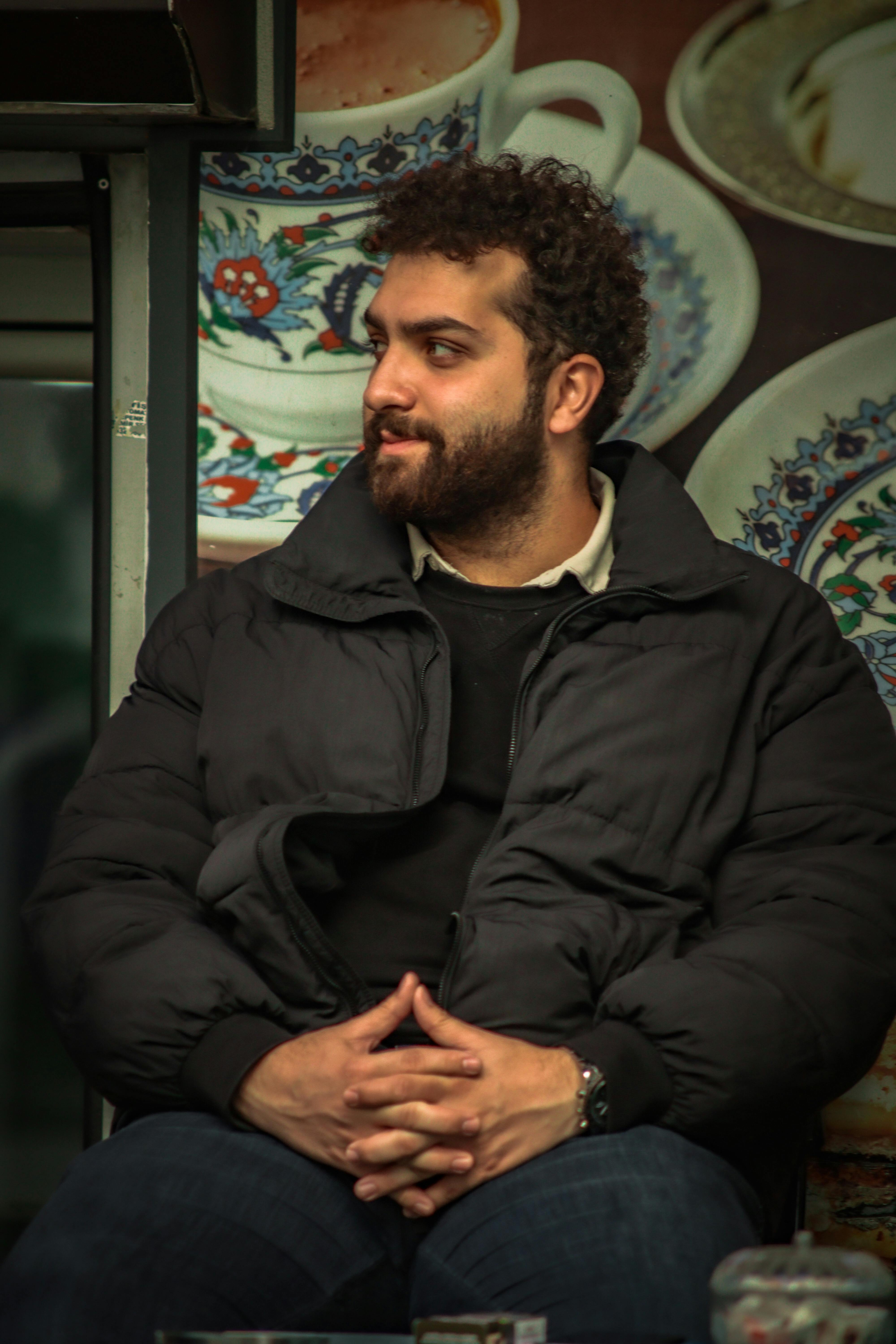 A cozy indoor portrait of a man in a black jacket, seated with patterned tea cups in the background.