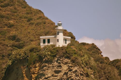 Free Beautiful lighthouse perched on a rugged cliff, surrounded by lush greenery under a clear sky. Stock Photo