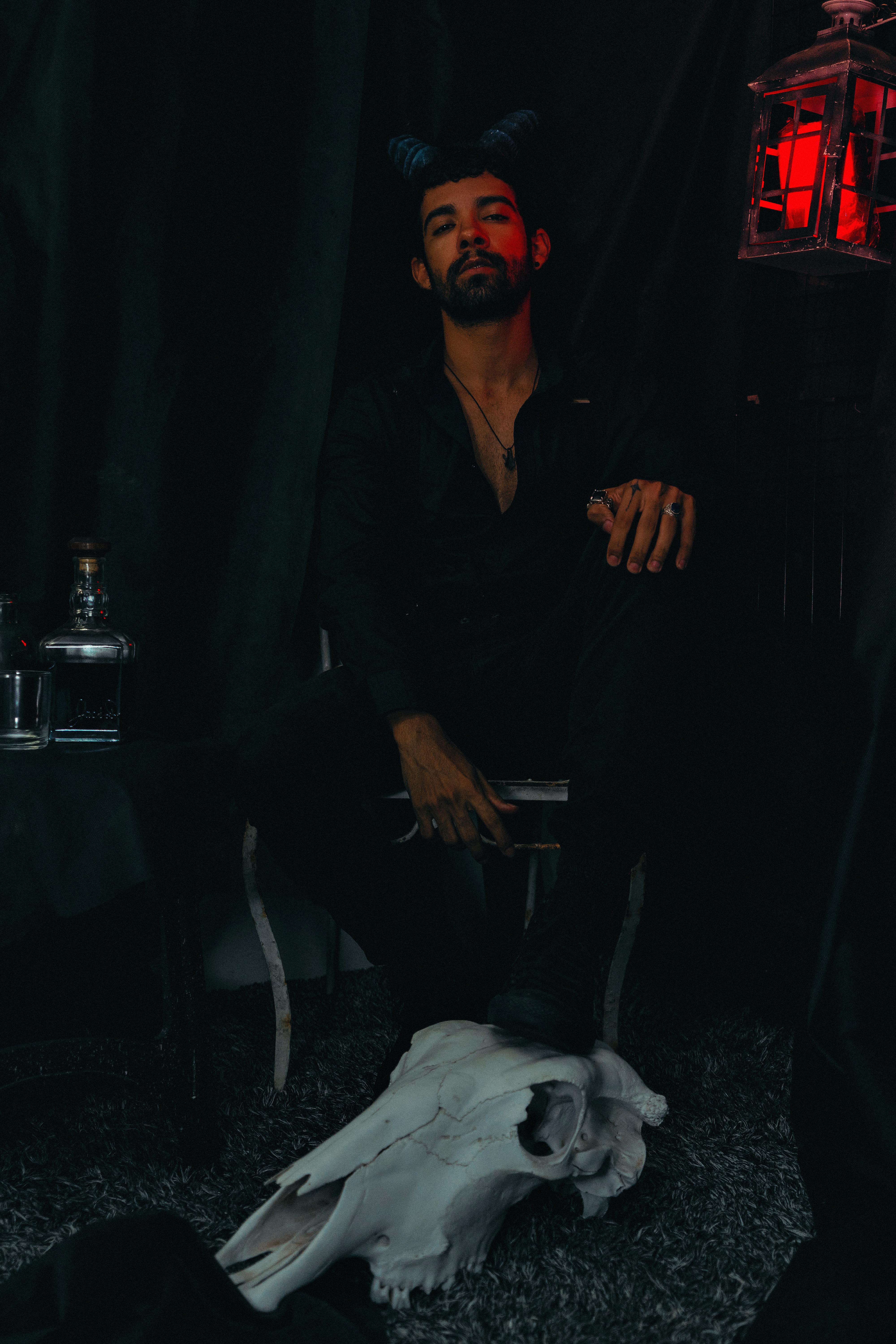 grátis Retrato com tema escuro de um homem com chifres sentado ao lado de uma caveira em iluminação ambiente. Foto profissional