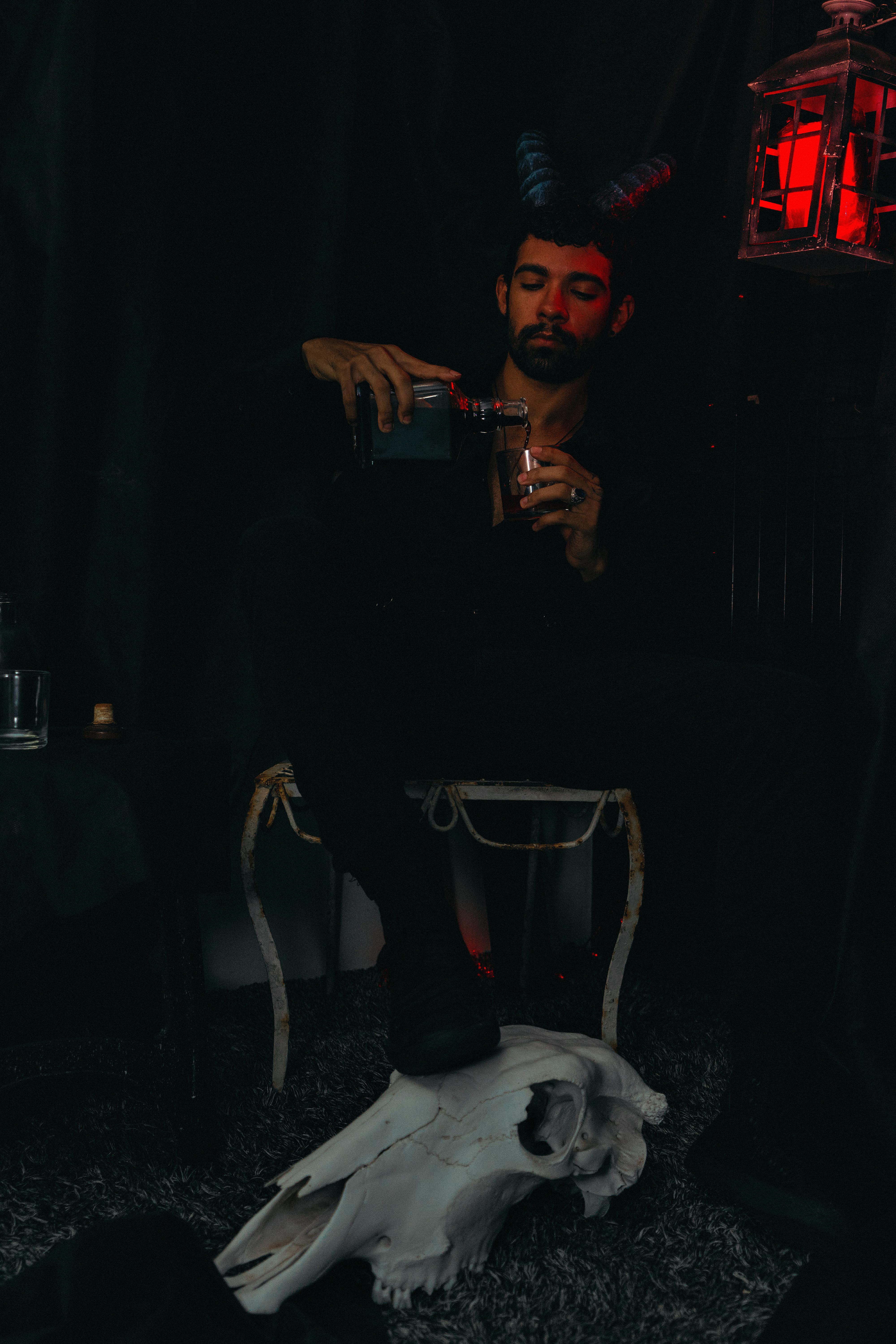 grátis Um homem com chifres está sentado em uma sala escura, servindo uma bebida, exalando uma vibração misteriosa e temperamental. Foto profissional