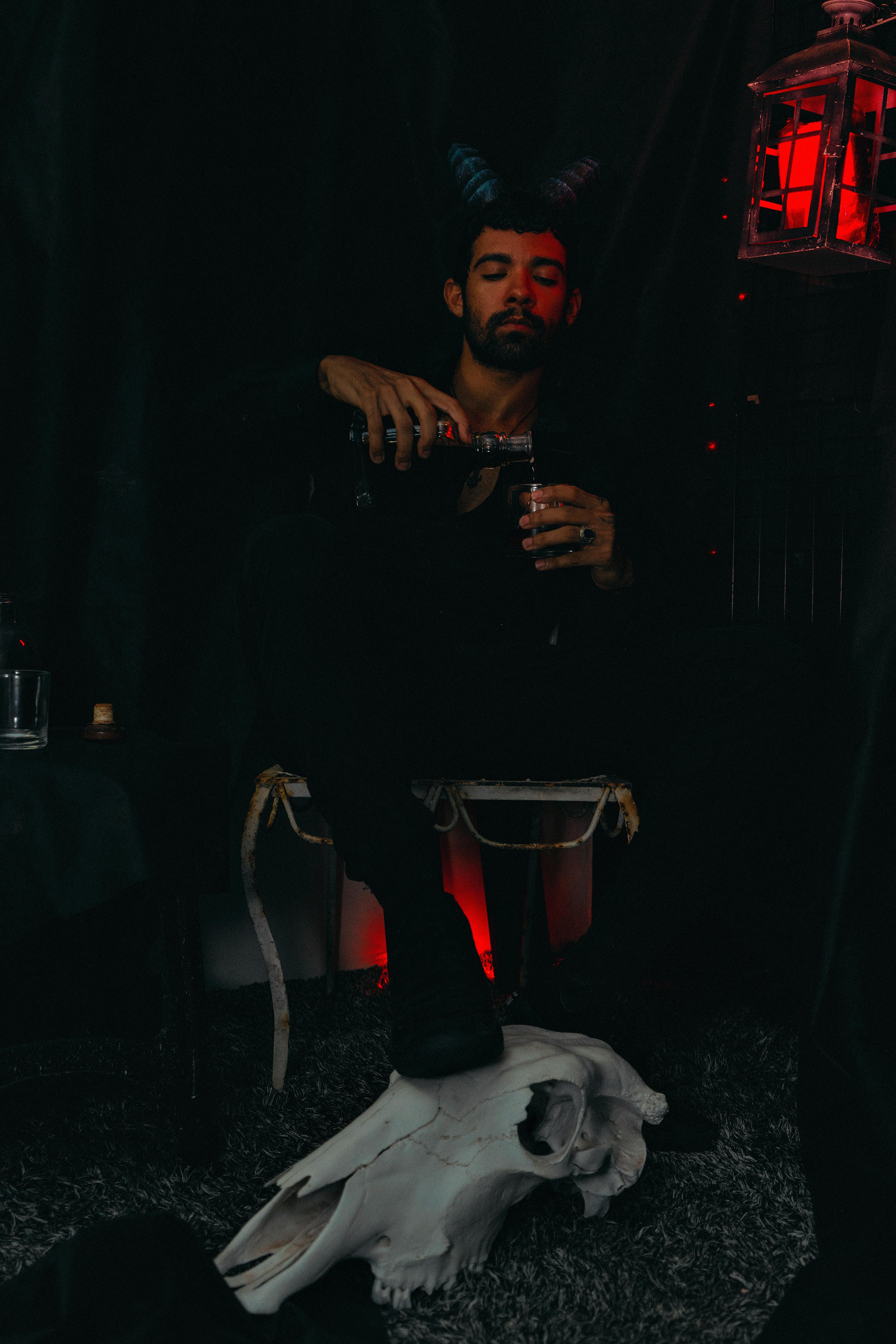 Man with horns pouring drink in a dark Halloween setup with skull and red lantern lighting.