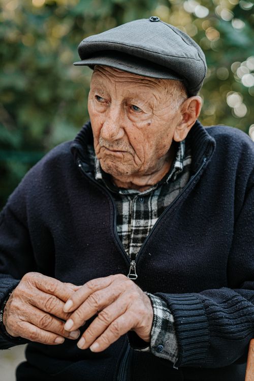 Free Portrait of an elderly man wearing a cap, sitting thoughtfully outdoors. Stock Photo