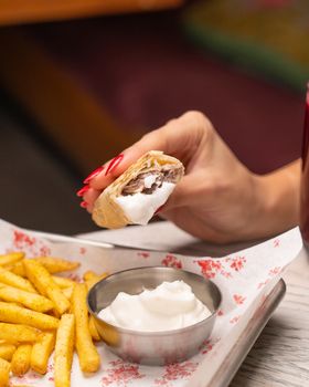 Close-up of hand holding tasty wrap with fries and sauce on a tray.