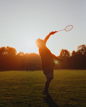 Silhouette of a person playing badminton in a grassy field at sunset, creating a dramatic scene.