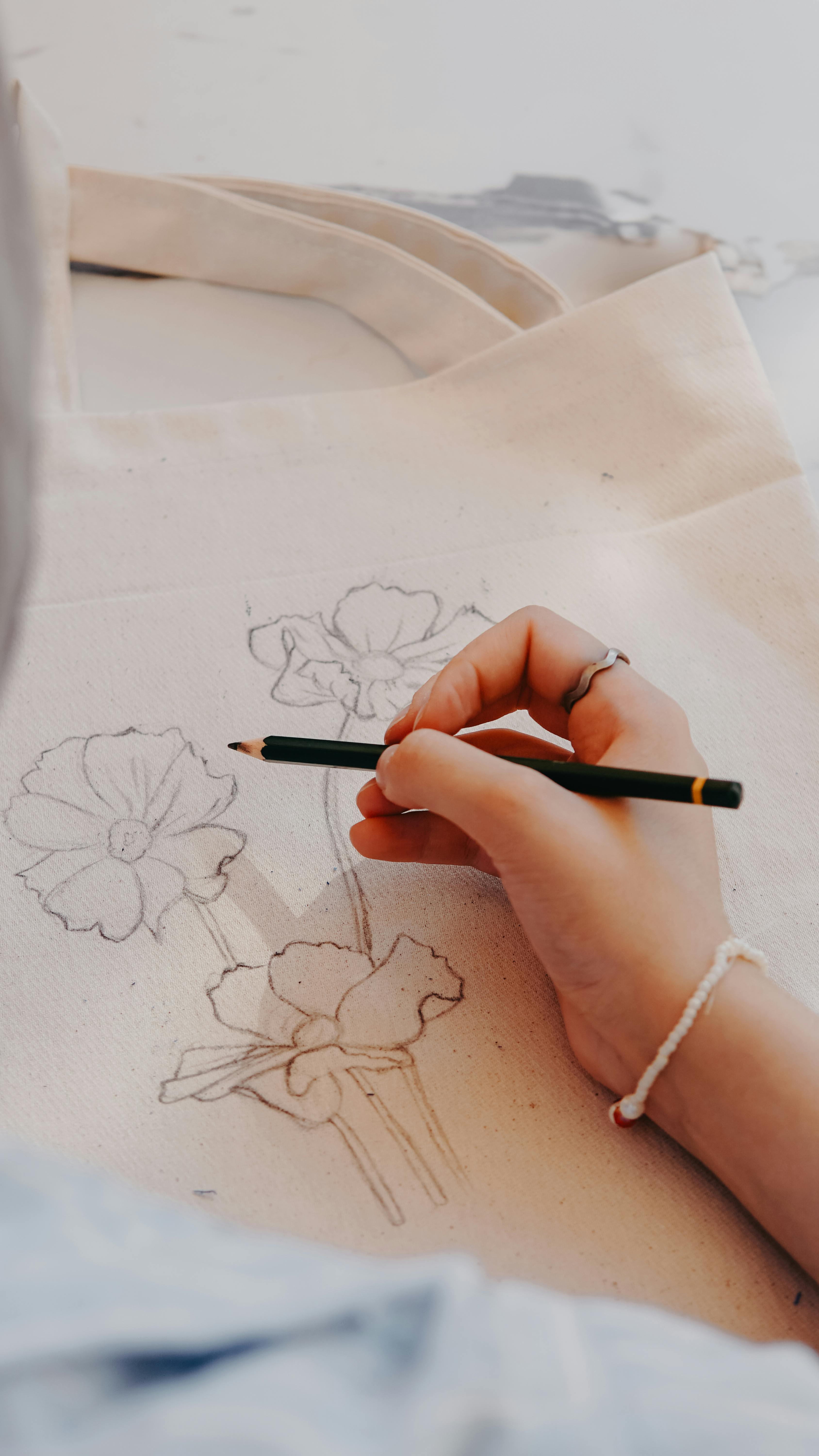 A close-up of an artist's hand sketching floral designs on a canvas tote bag.