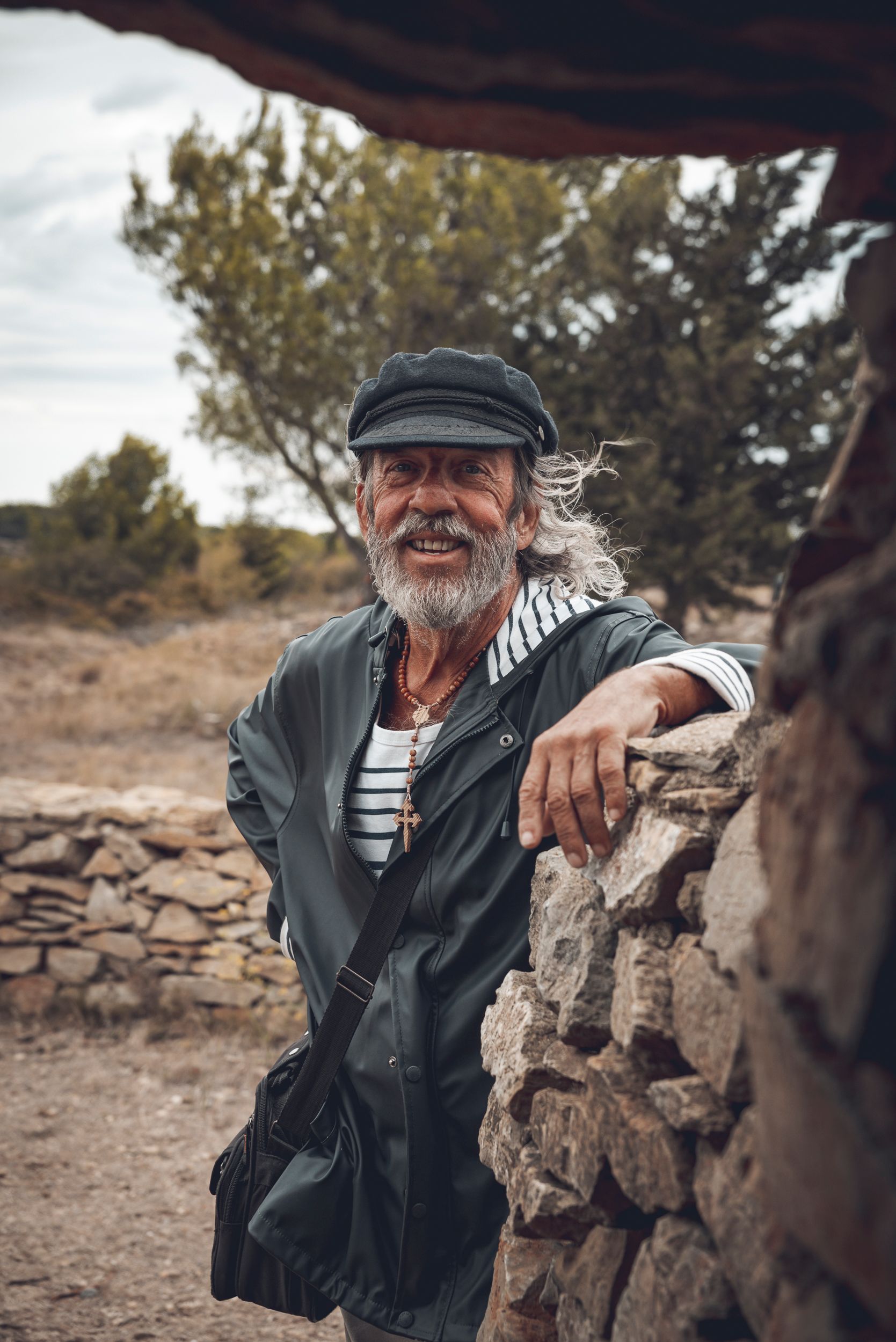 Elderly sailor with beard and cap leaning on stone wall outdoors, smiling warmly.