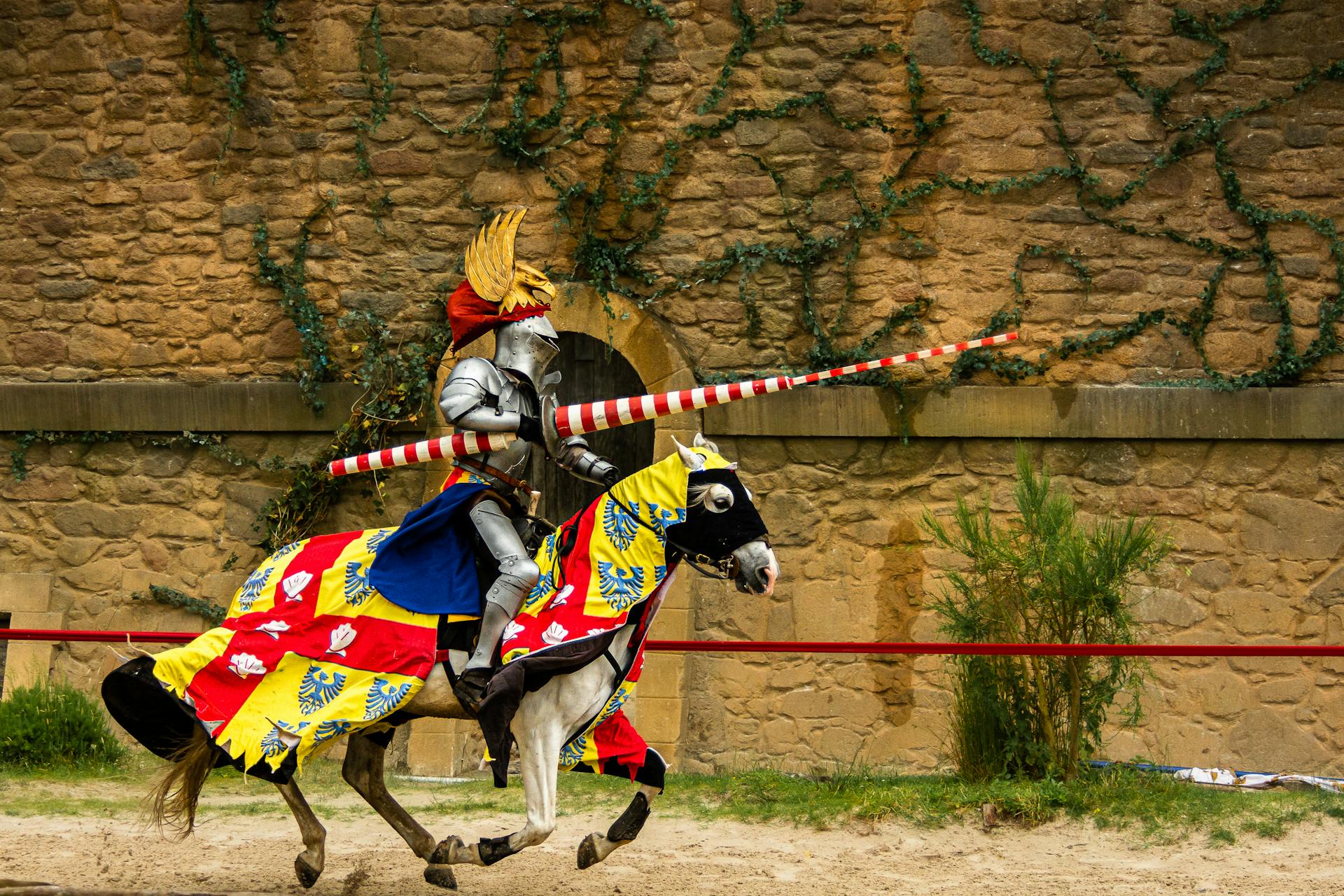 A medieval knight in full armor jousting on a decorated horse against a rustic stone wall backdrop.