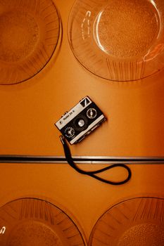 Retro analog camera with a strap on a vibrant orange surface surrounded by clear textured plates.