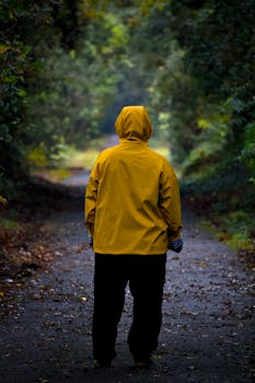 A person in a yellow coat walks alone on a forest path surrounded by lush greenery.