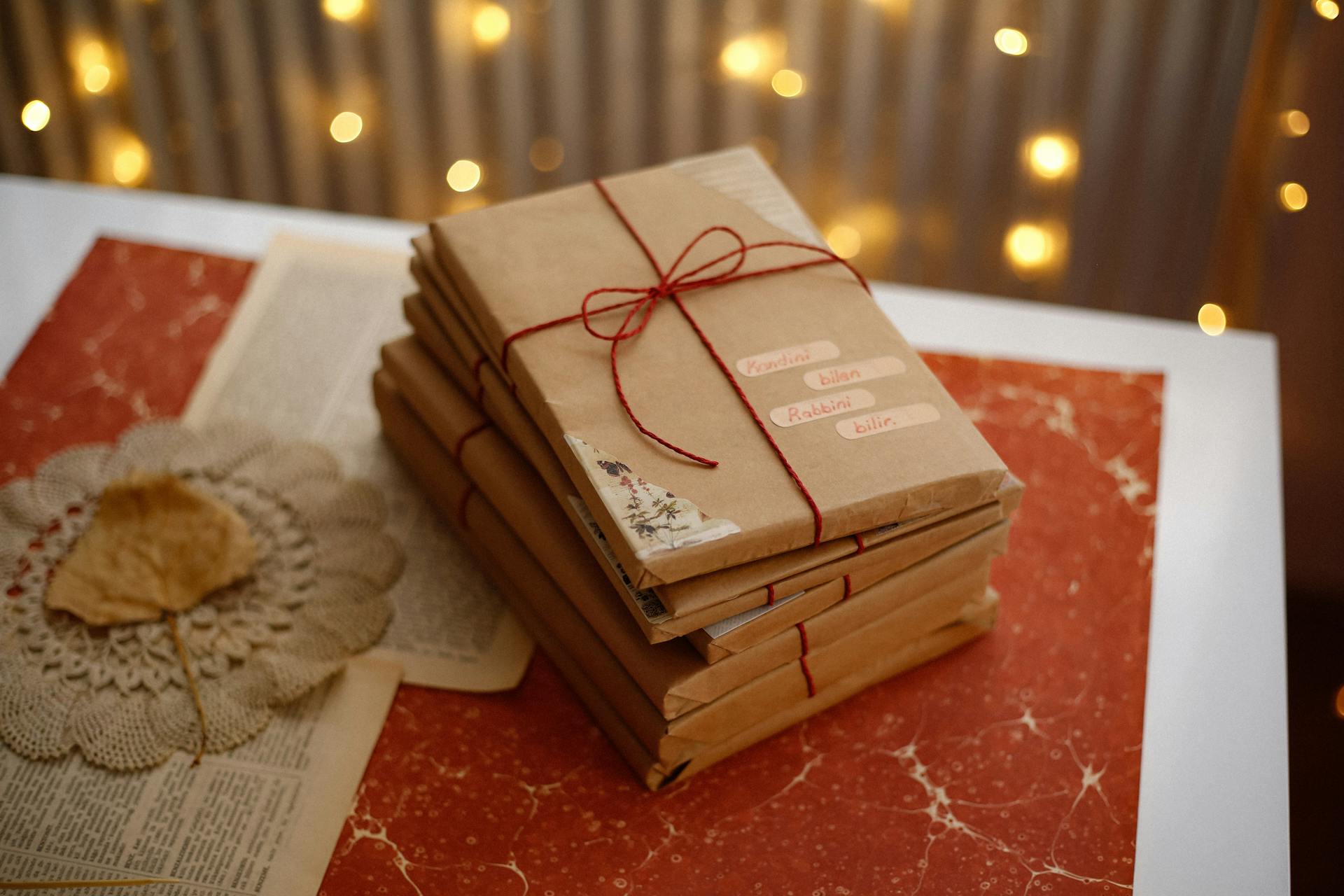 A stack of wrapped books tied with red string, set against a warm, festive backdrop with string lights.