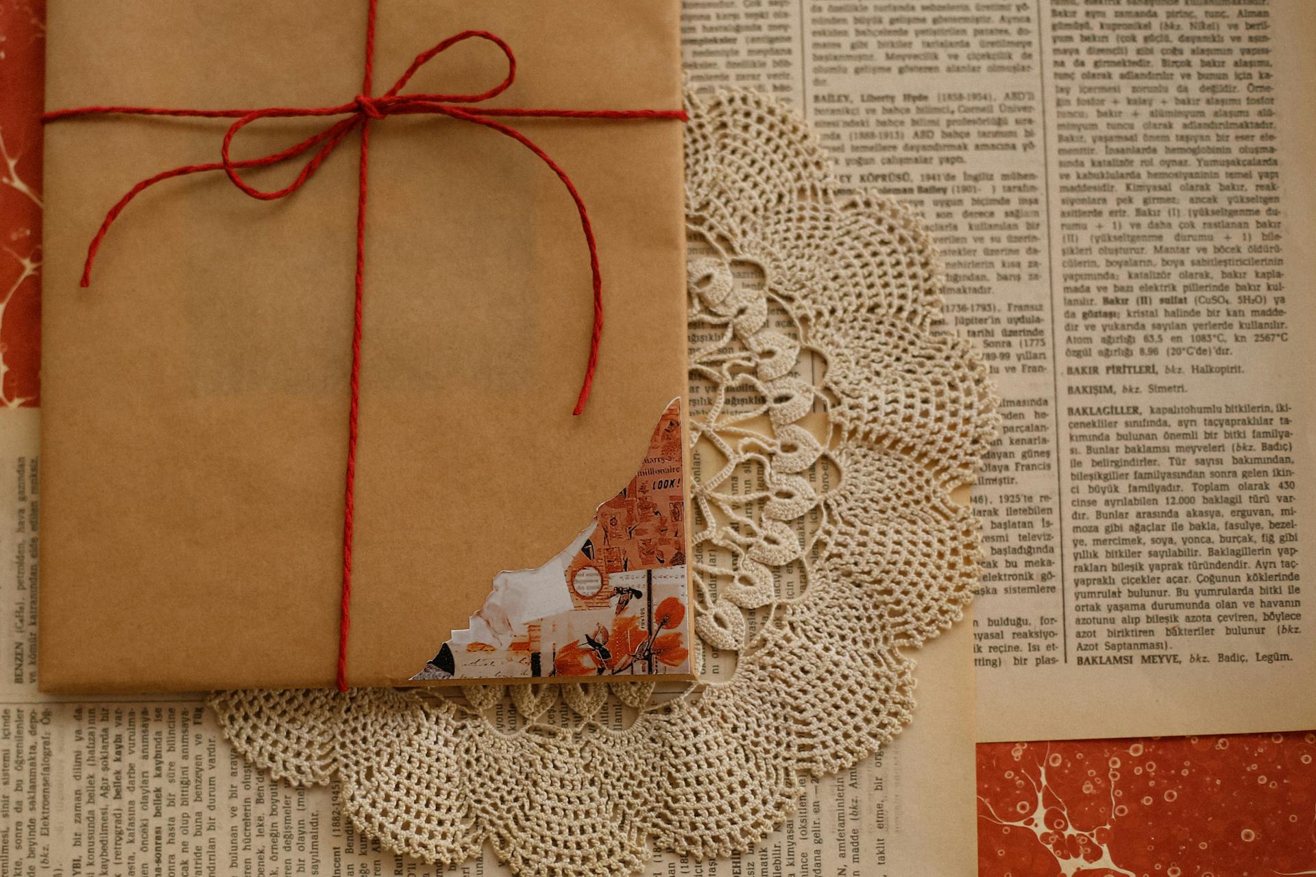 A vintage-styled wrapped gift with brown paper and red string on lace doily, atop old newspapers.