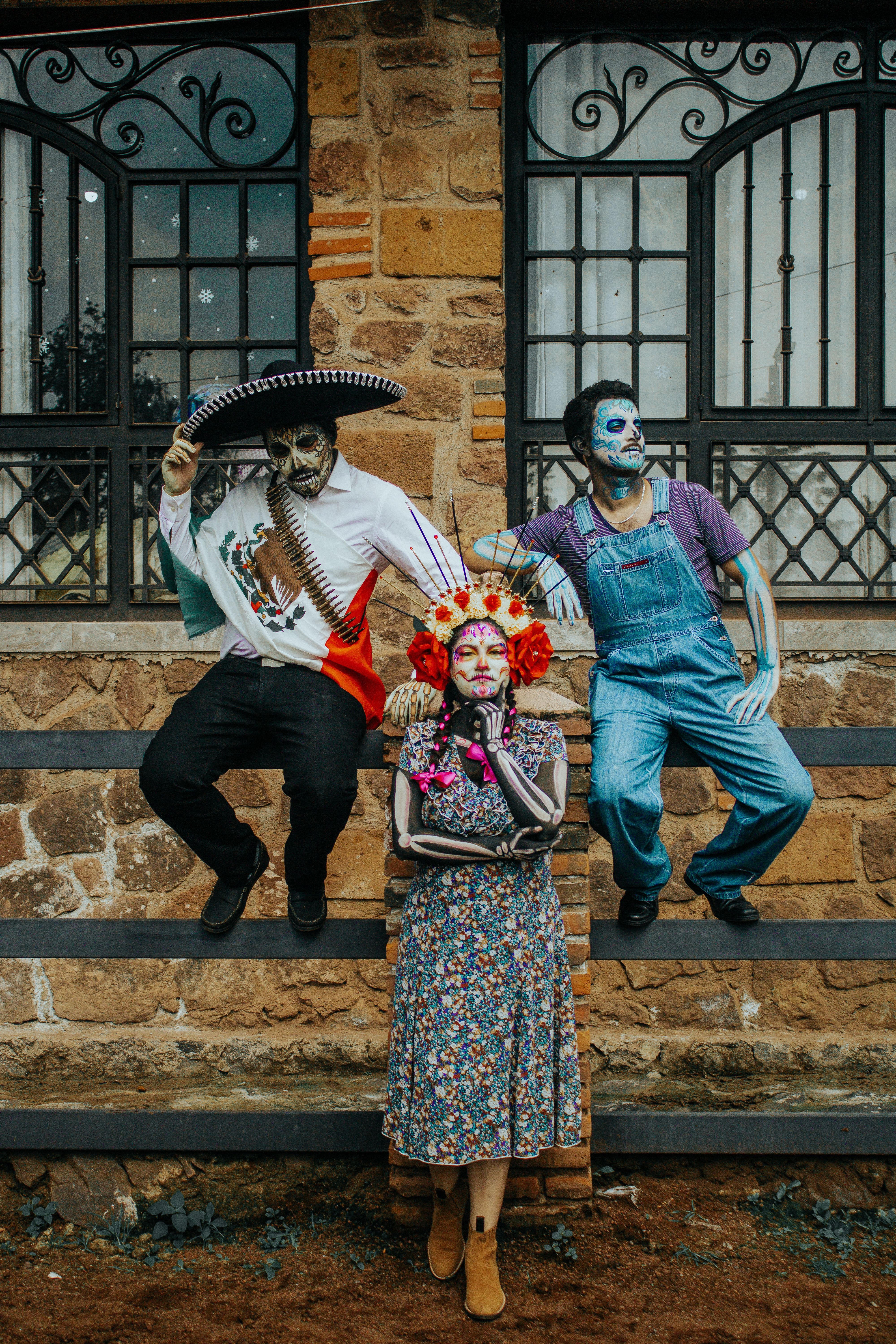 Three people dressed in traditional Day of the Dead costumes pose against a rustic background.