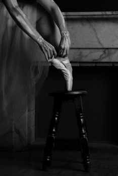Black and white photo of a ballet dancer adjusting pointe shoes on a stool elegantly.