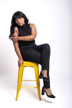Confident woman in black attire posing on a yellow stool against a white background.