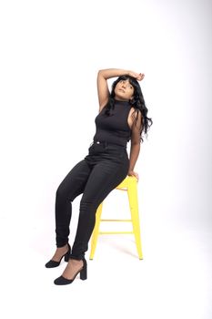 Fashionable woman in black outfit posing confidently on a bright yellow stool in a studio environment.