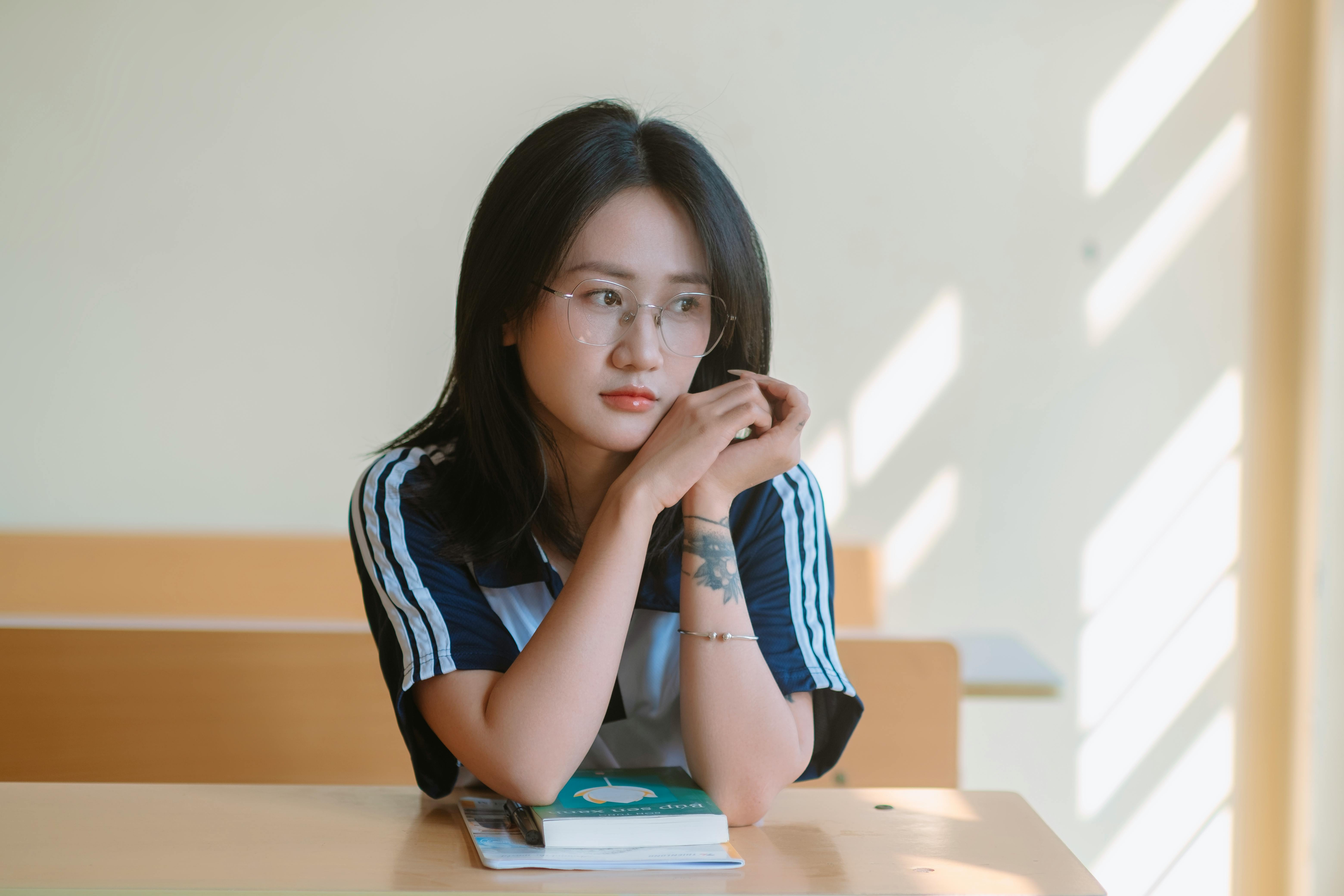 무료 교실에 있는 한 젊은 여성이 햇살이 쏟아지는 것을 생각에 잠긴 듯 바깥을 바라보고 있습니다. 스톡 사진