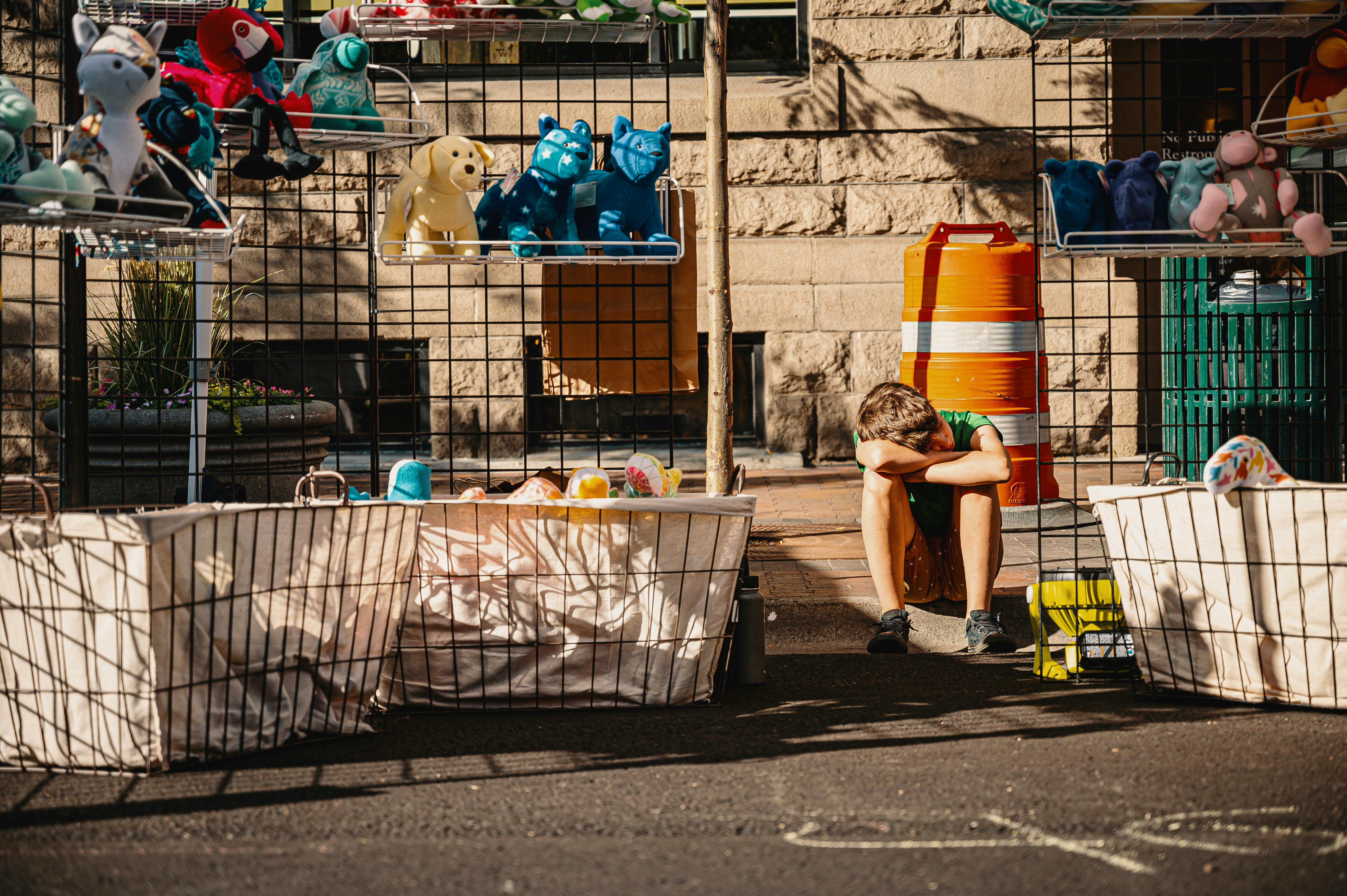 boy crouching and resting head on hands and knees between shelf of plush toys