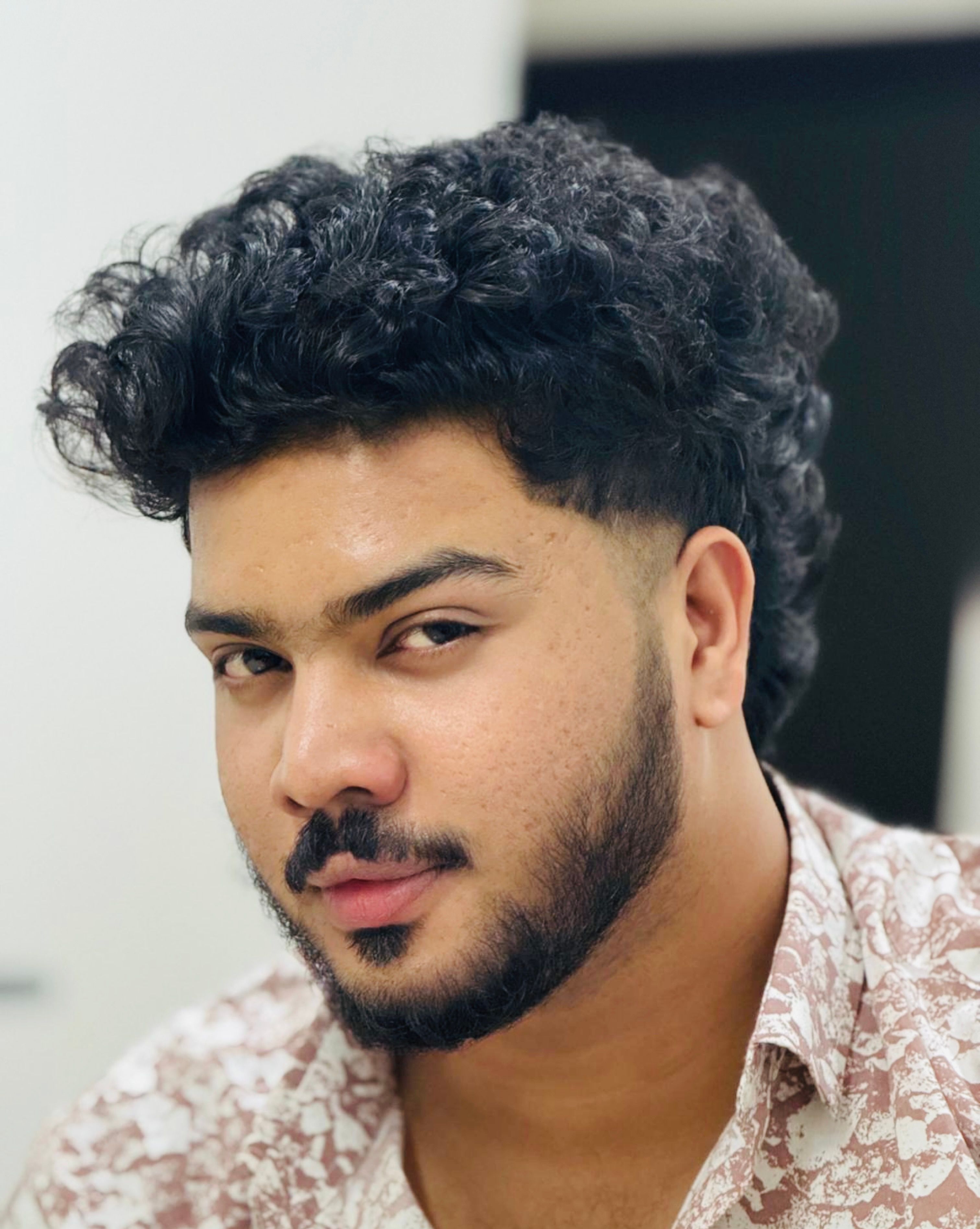 Close-up portrait of a young man with curly hair and a beard, looking confidently at the camera.