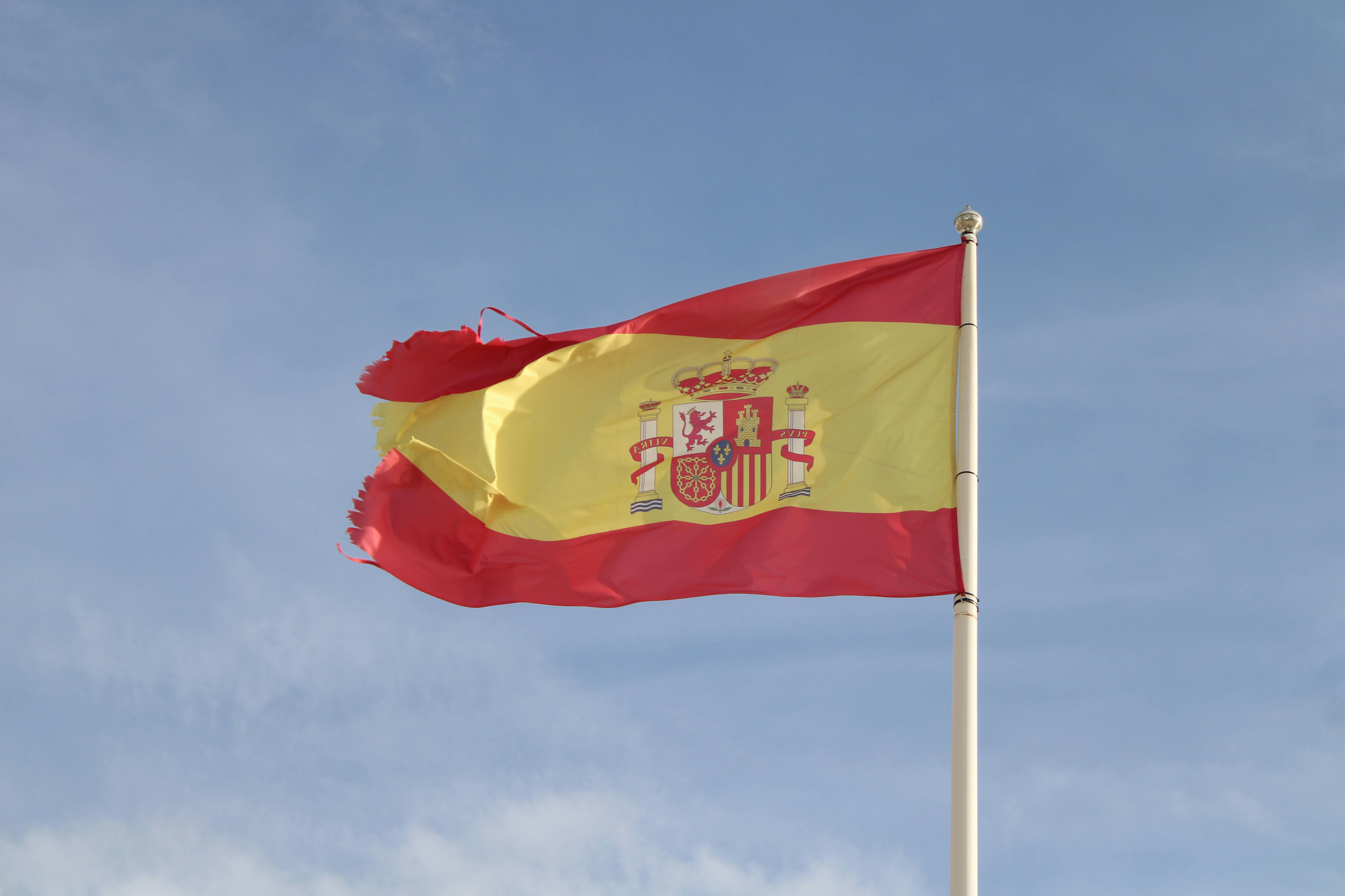 Spanish flag waving on a tall flagpole against a clear blue sky in El Puerto de Santa María, España.