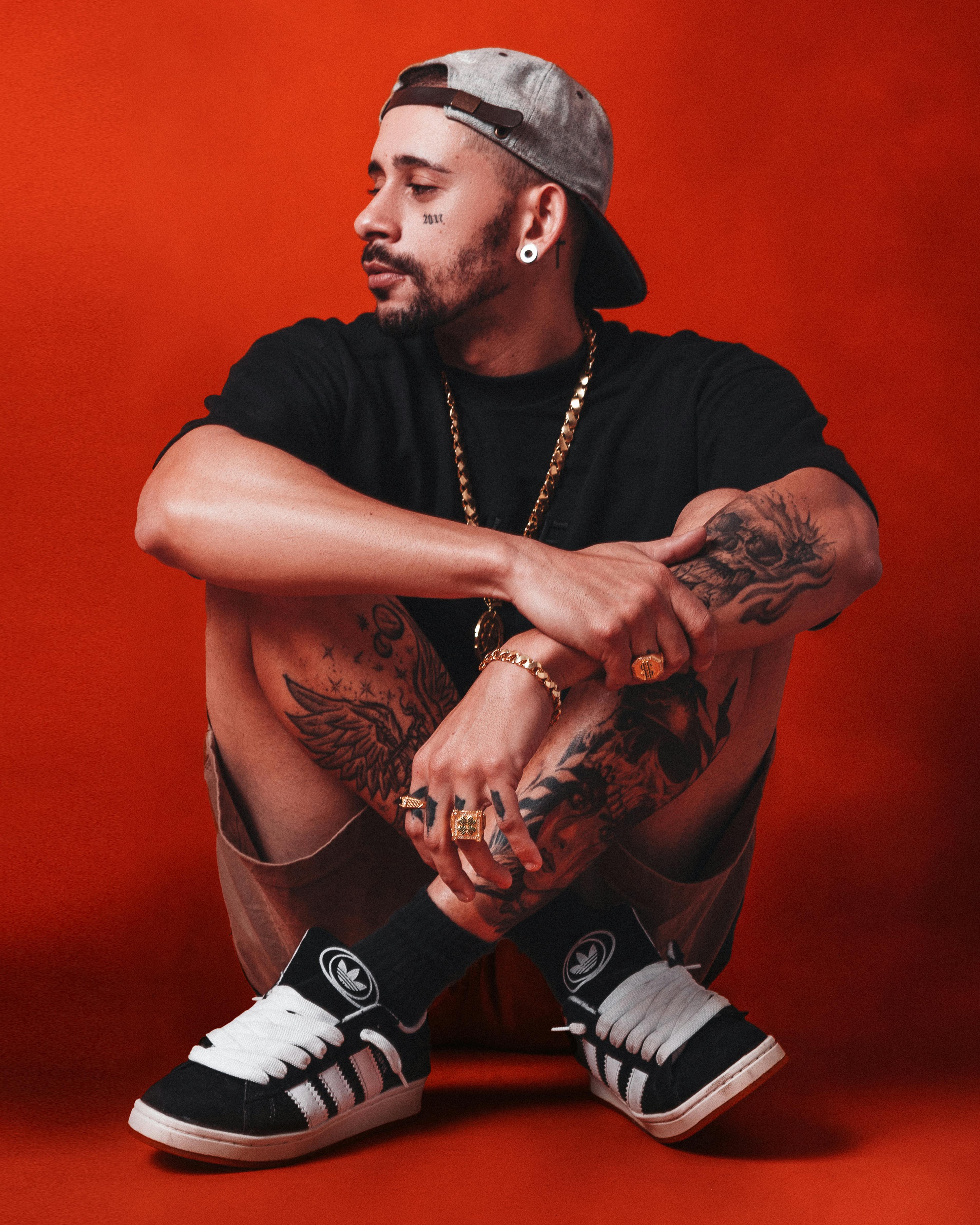 A fashionable young man with tattoos seated on a red background, exuding urban style.