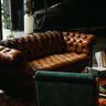 A classic, tufted brown leather Chesterfield sofa sits in an industrial-style room with moody lighting.