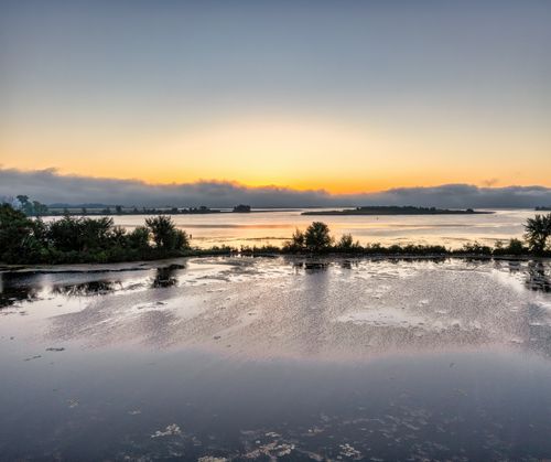 Free Peaceful sunrise over a calm lake with reflective waters and lush trees, offering a serene landscape view. Stock Photo