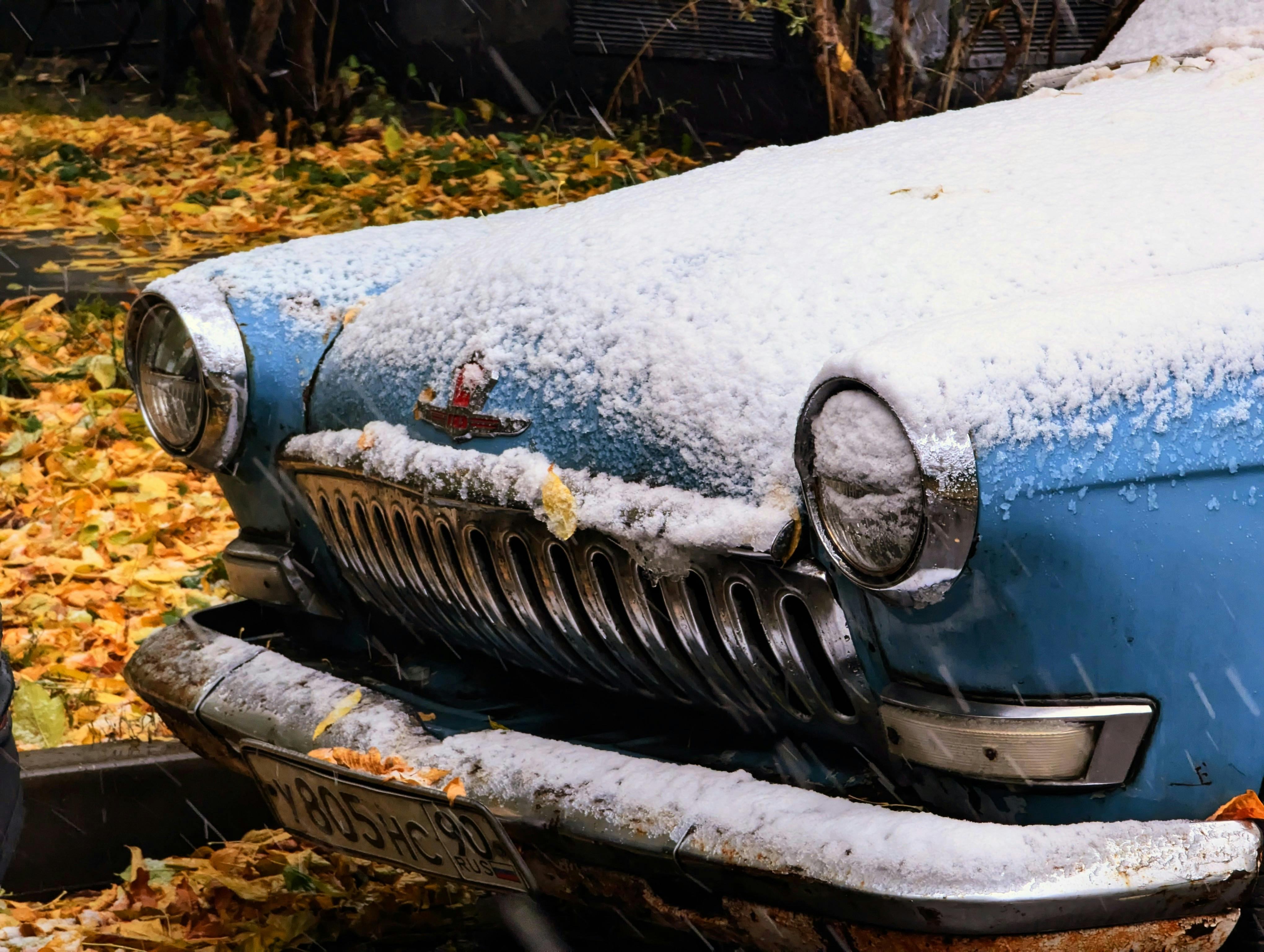 Close-up of a classic blue car dusted with snow, contrasted by fallen autumn leaves.
