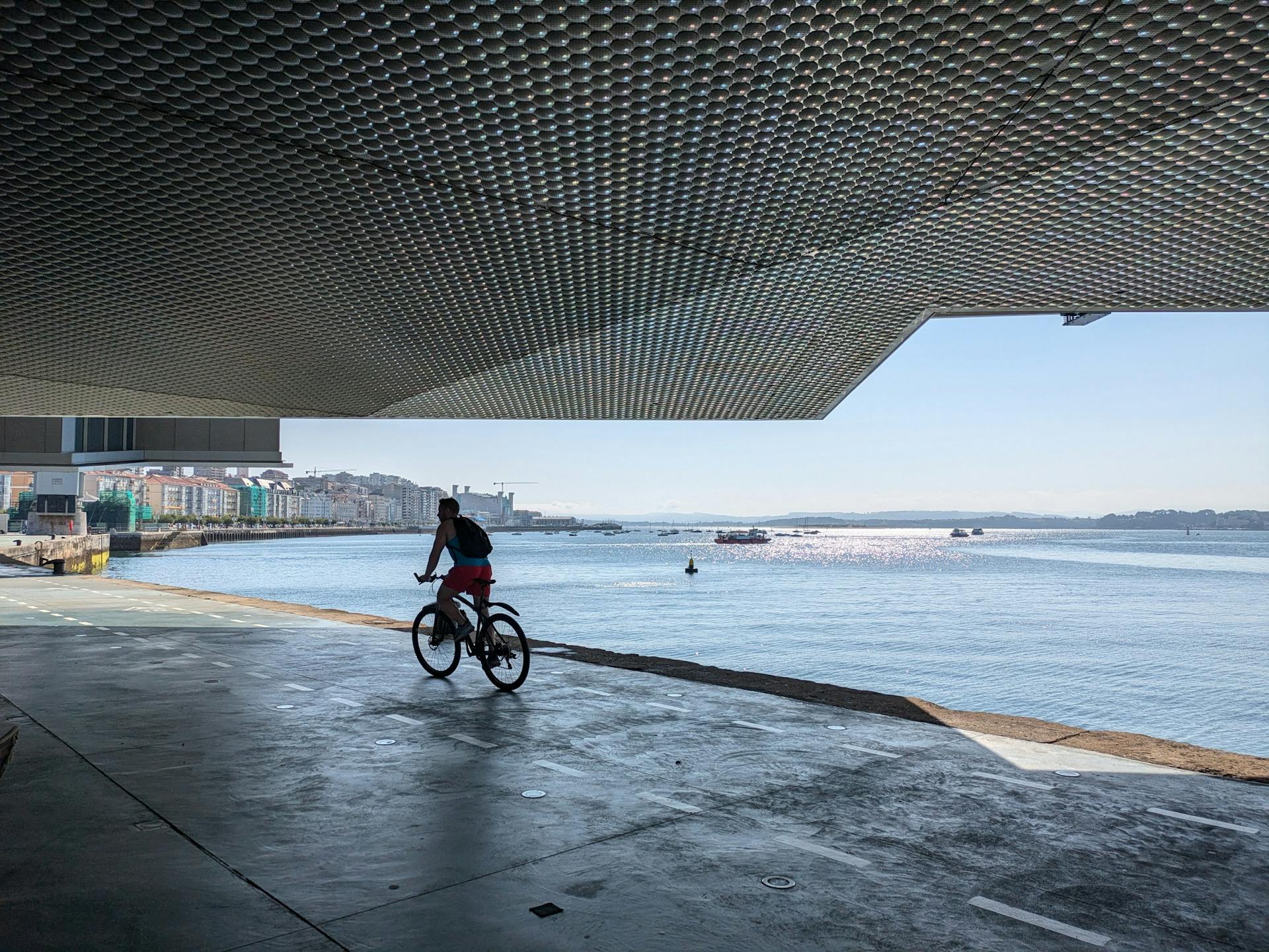 A cyclist rides along the coast under a modern structure in Santander, Spain.