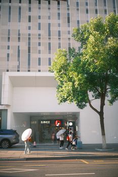 Urban street view with people walking, carrying umbrellas and a tree outside a modern building.