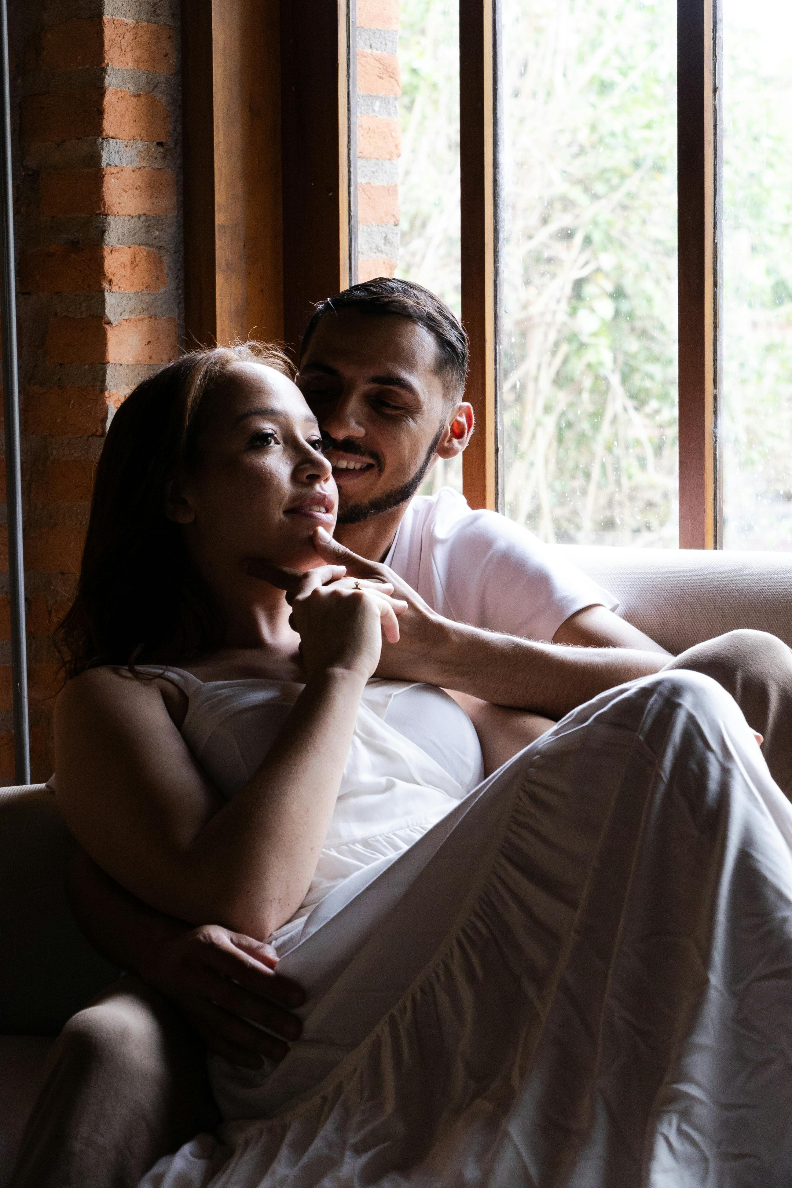 Lovers in tender embrace by rain-streaked window, soft morning light filtering through, peaceful intimacy
