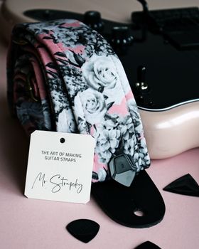 Black electric guitar with floral strap and picks perfectly arranged on a pastel surface.