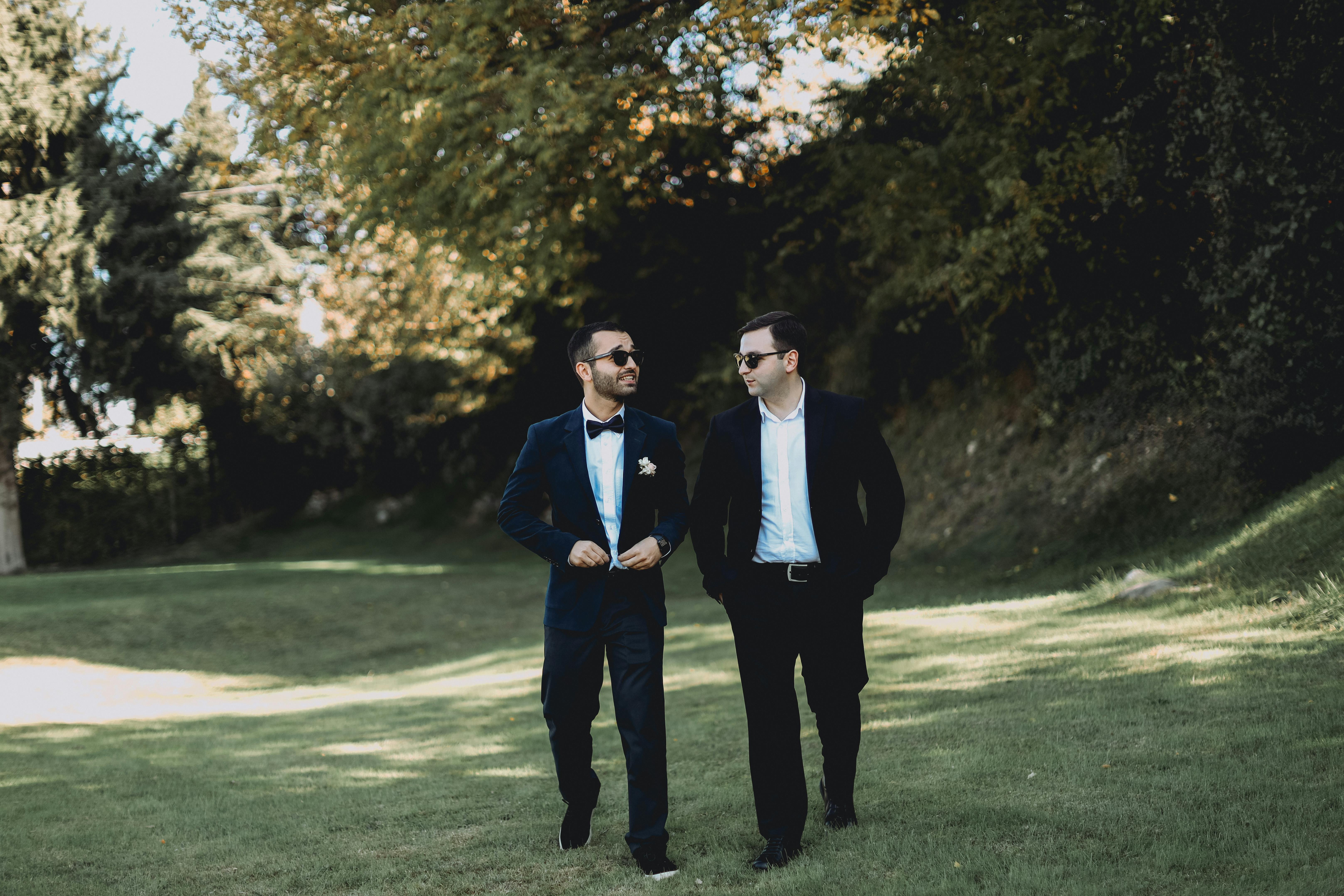 Two men in formal suits walking on a grassy area under trees, exuding confidence and style.