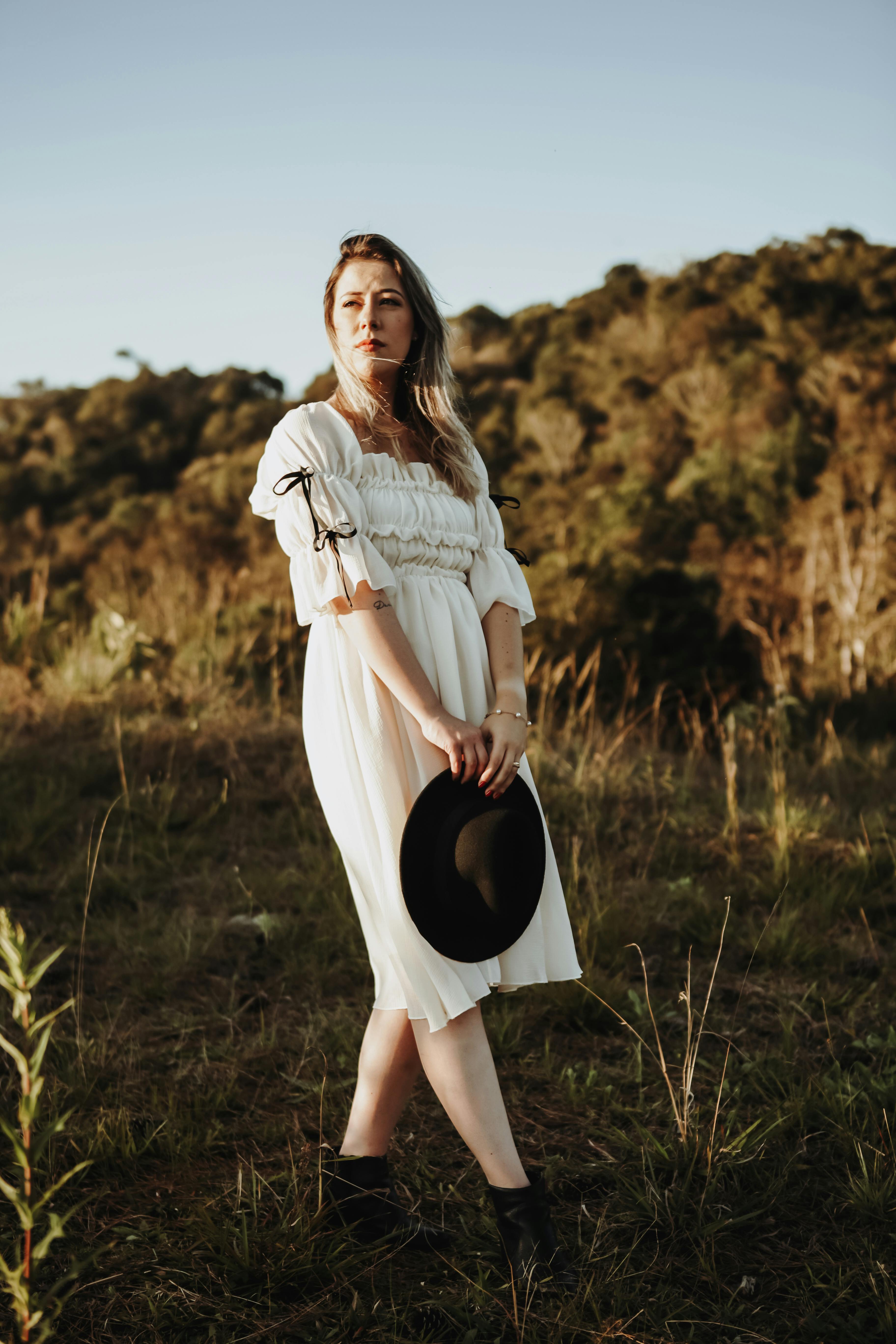 Portrait of a young woman in a white dress holding a hat outdoors in a sunny meadow.